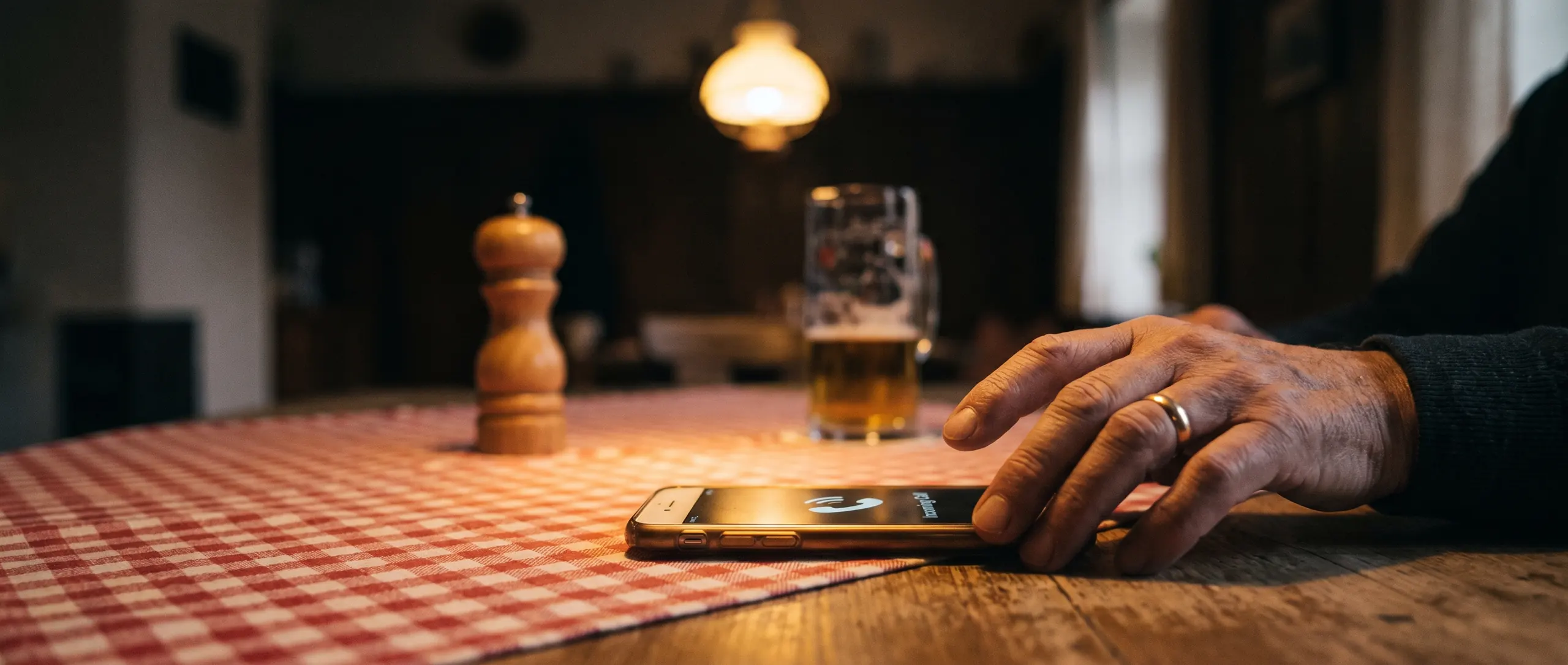 Die Hand eines älteren Mannes auf einem klingelnden Handy, das auf dem rot karierten Tuch eines Gasthaustischs liegt, daneben ein Krug Bier.