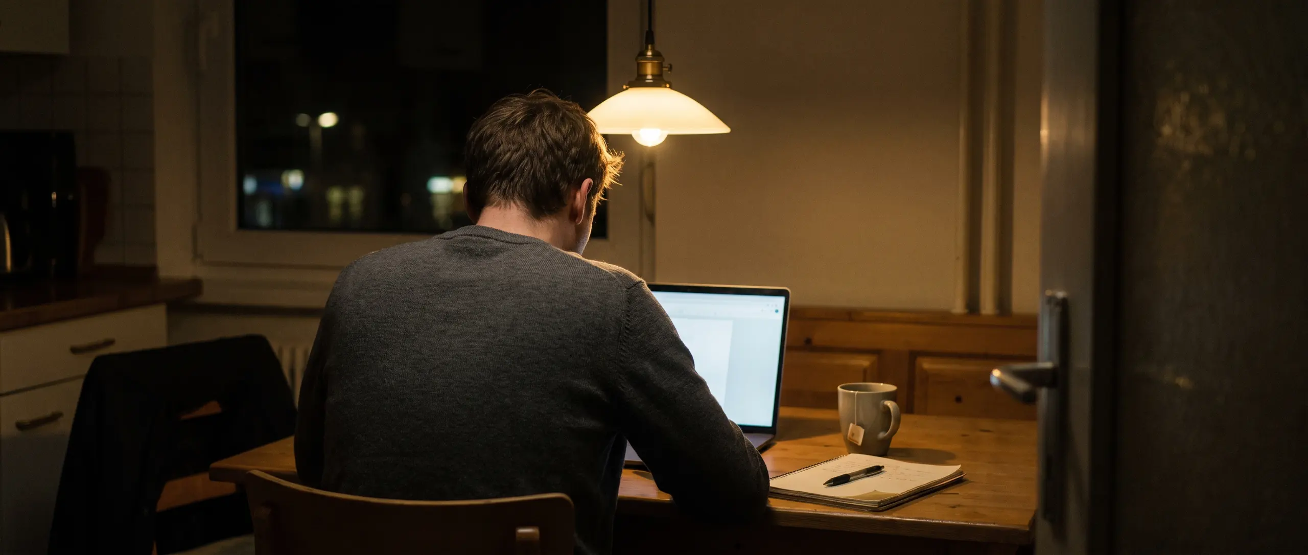 Ein Mann sitzt abends von hinten gesehen am Küchentisch vor einem Laptop, daneben ein Notizbuch und eine Tasse.